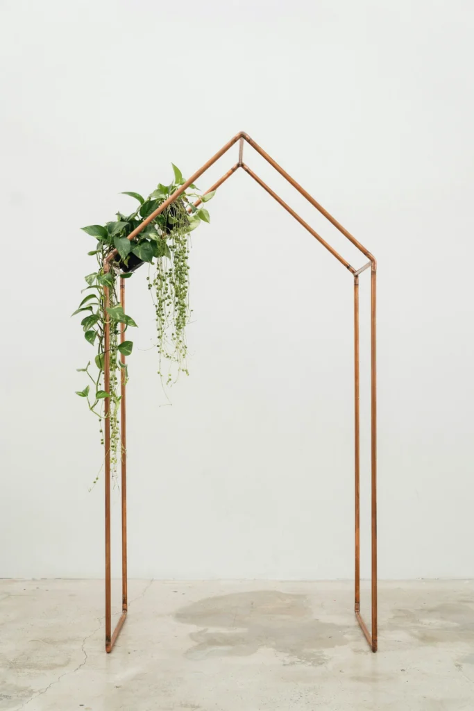 A clean, angular copper hexagonal arch with only trailing greenery — no flowers — against a white wall or open sky; simple, architectural, and strikingly modern