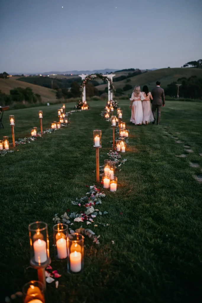 A winding grass pathway between the home and the ceremony space, lined with alternating heights of glass lanterns holding white pillar candles, at dusk — guests walking toward the glow