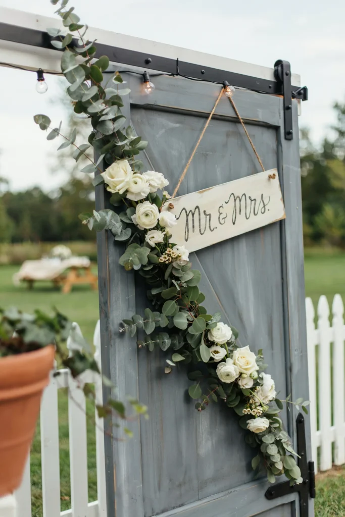 A reclaimed barn door panel propped against a fence or wall in a backyard, styled with hanging eucalyptus, white blooms, and a wooden "Mr. & Mrs." sign — used as a photo booth backdrop