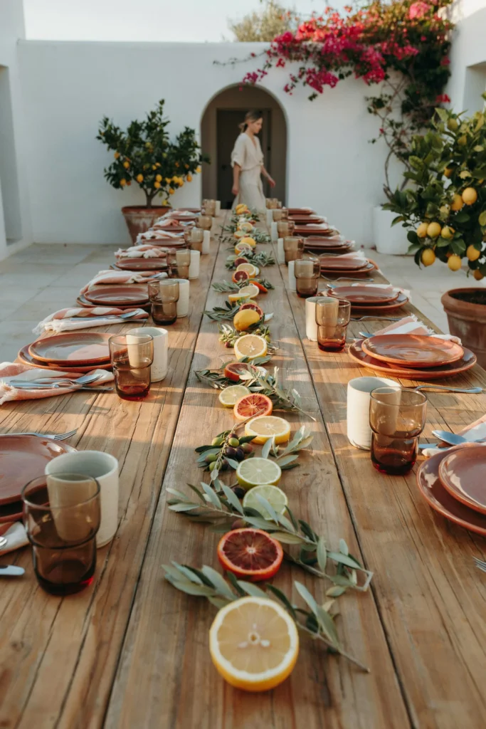 Long outdoor table with potted lemon trees at each end, terracotta charger plates, citrus slices scattered among olive branch centerpieces, striped linen napkins in terracotta and cream, and warm golden light
