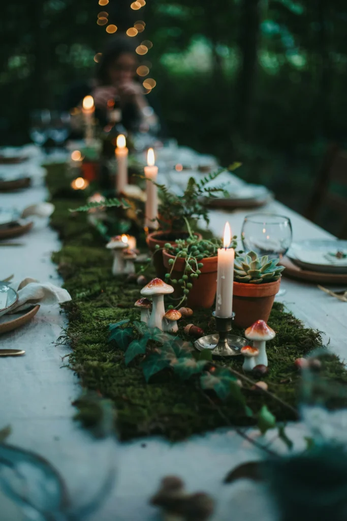  Low tablescape with a moss runner down the center, small terracotta pots of ferns and succulents, scattered mushroom figurines, soft white taper candles, and trailing ivy over the table edge