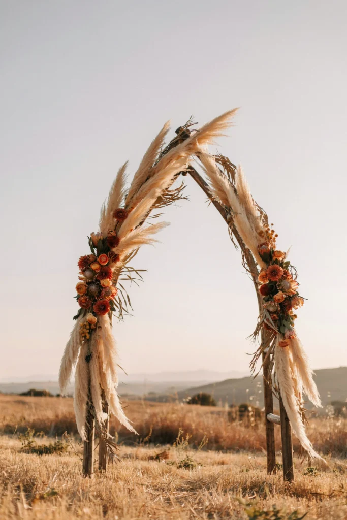 Tall, organic ceremony arch made of white pampas grass, dried orange and terracotta florals, and trailing dried palm fronds — warm and textural — set against a golden-hour sky