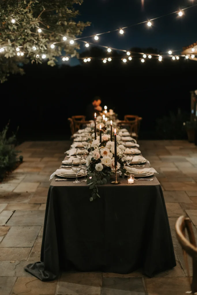 An elegantly dressed backyard at night — long black linen tablecloths, towering white floral centerpieces with black foliage, black taper candles, crystal glassware, and twinkling overhead lights — sophisticated and unexpected