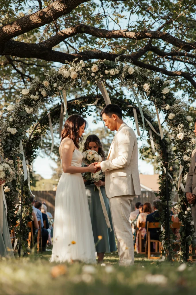 A couple exchanging vows under a large backyard tree draped with hanging flowers and greenery, two witnesses standing close, warm filtered light through the leaves — the simplest and most beautiful ceremony setup possible