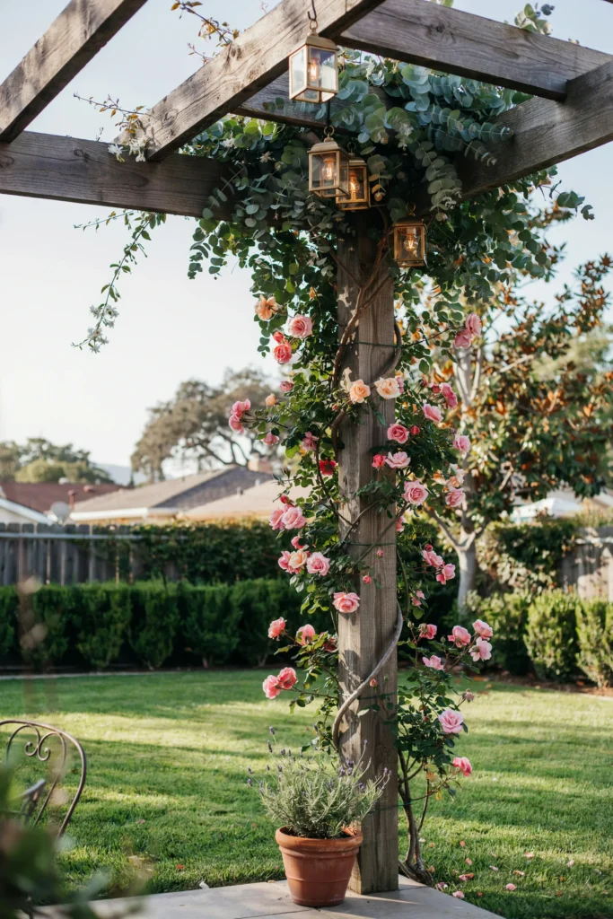 A small backyard with a towering floral and greenery installation on a trellis or pergola column — climbing roses, hanging eucalyptus, and small lanterns — drawing the eye upward and making the space feel larger
