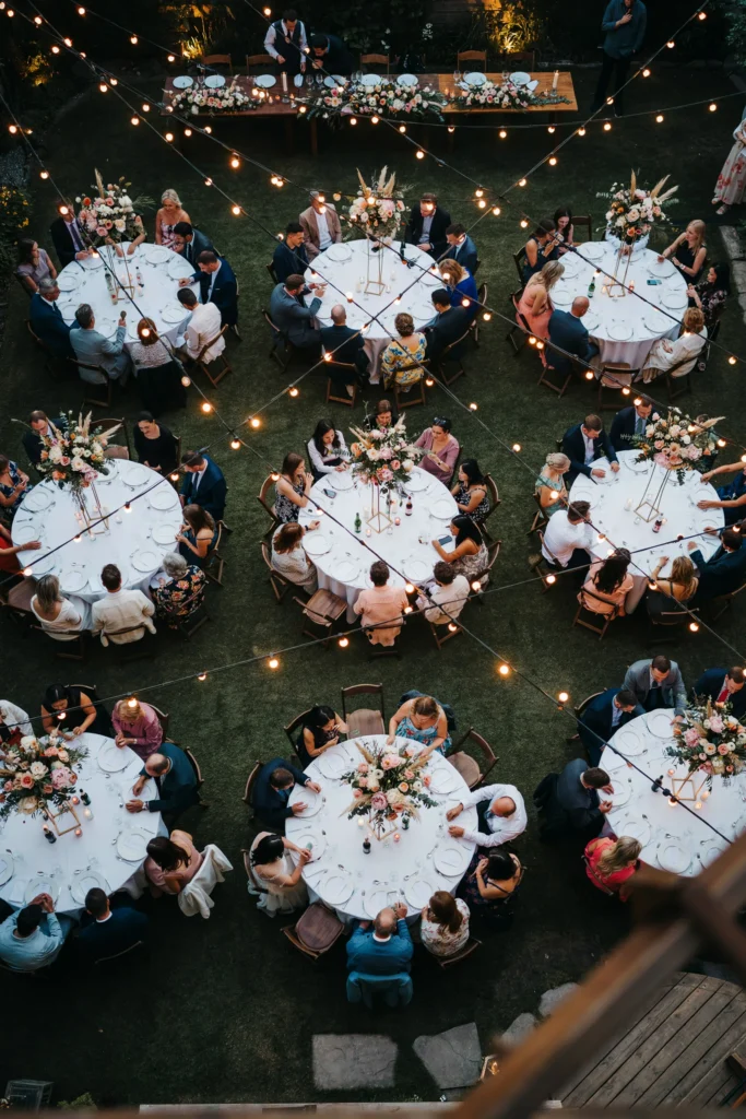  Overhead view of a backyard reception with dense Edison bulb string lights creating a glowing canopy above round tables set with white linens and tall floral centerpieces at dusk