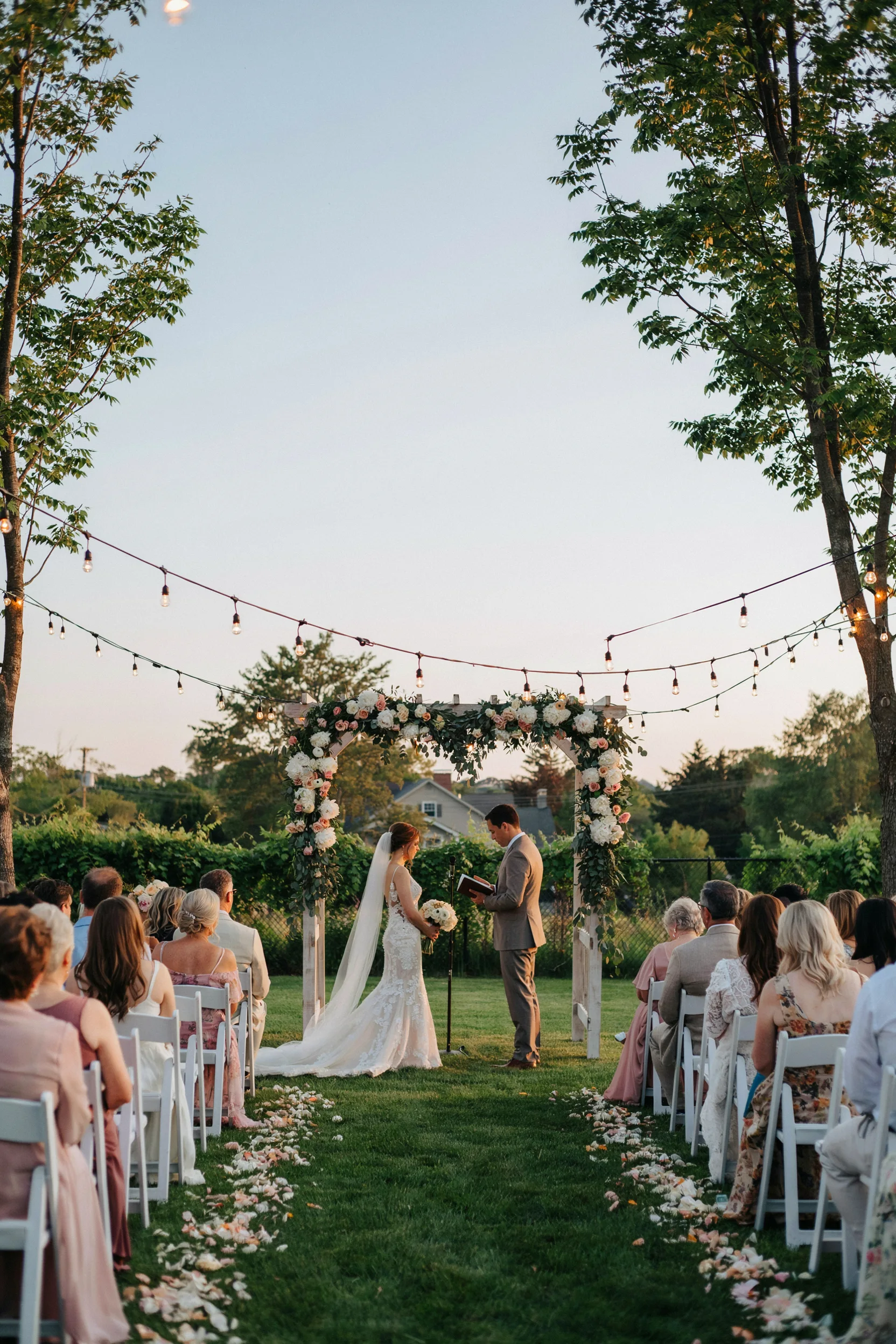 A warmly lit backyard wedding at golden hour — string lights draped overhead, a wooden arch wrapped in white florals and greenery, guests seated in white chairs on a lush lawn.