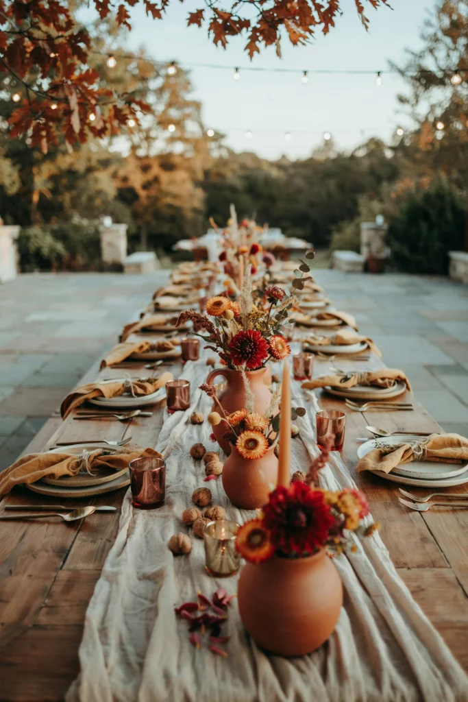 Outdoor autumn reception table with terracotta ceramic vessels, dried orange and rust florals, deep burgundy dahlias, scattered whole walnuts, and a neutral linen runner — warm and harvest-rich