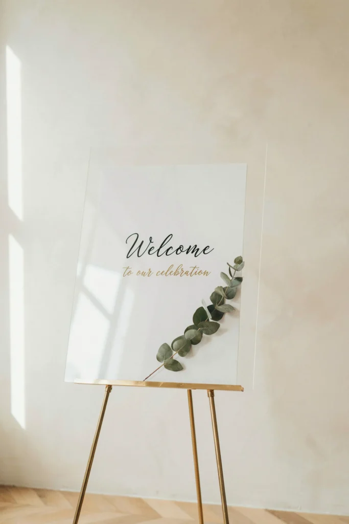  A clear acrylic welcome sign on a simple gold easel, with elegant script in black or gold, framed by a single sprig of eucalyptus — no table, no fuss, just one clean moment at the entrance