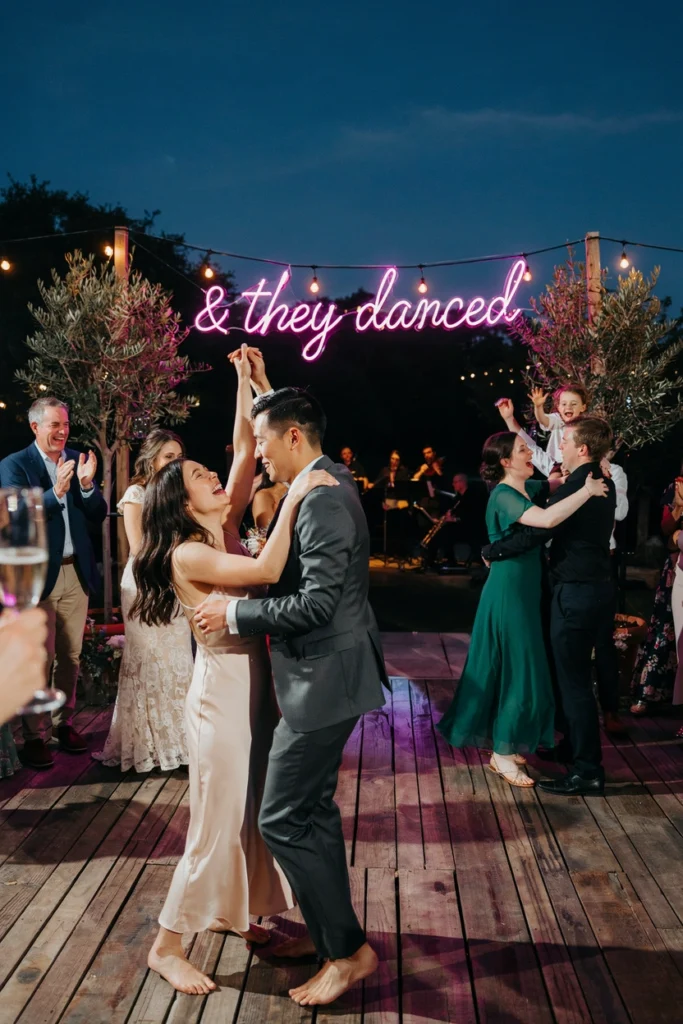 A custom neon sign reading "& they danced" hung above an outdoor dance floor at night — the warm pink glow against a dark sky reflecting off the faces of dancing guests