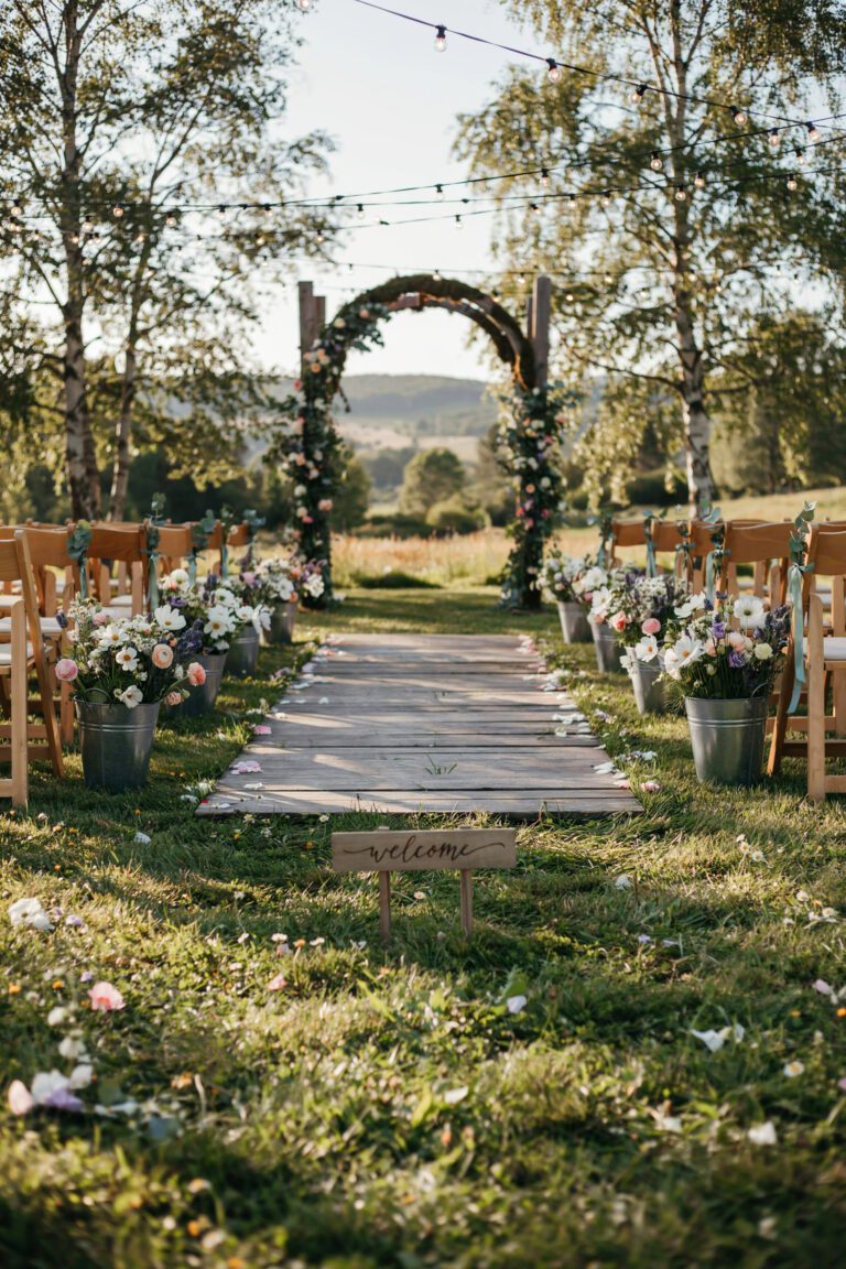 A sun-dappled outdoor ceremony space draped in soft string lights and trailing greenery, with a moss-covered arch framing the altar and wildflower arrangements lining the aisle