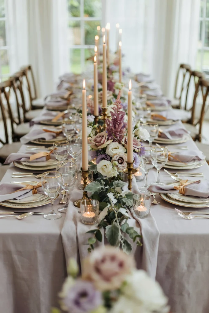 A richly layered table with soft lavender linen, white and lavender roses, purple astilbe, cascading silver leaves, tall gold candelabra, and ornate place settings with lavender napkins tied with gold ribbon.