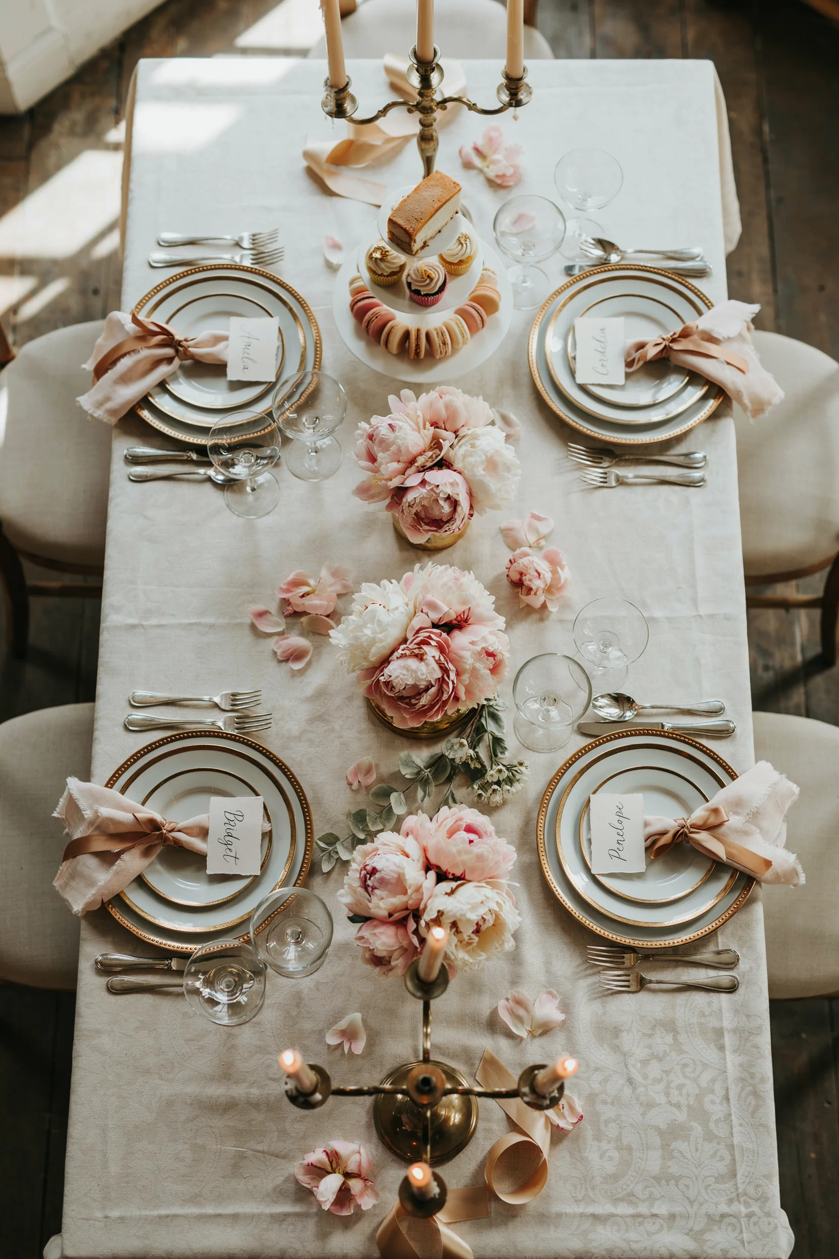 A luxurious overhead shot of an elegant Bridgerton-inspired bridal shower table setting with blush peonies, gold candlesticks, tiered dessert stand, and soft, romantic lighting.