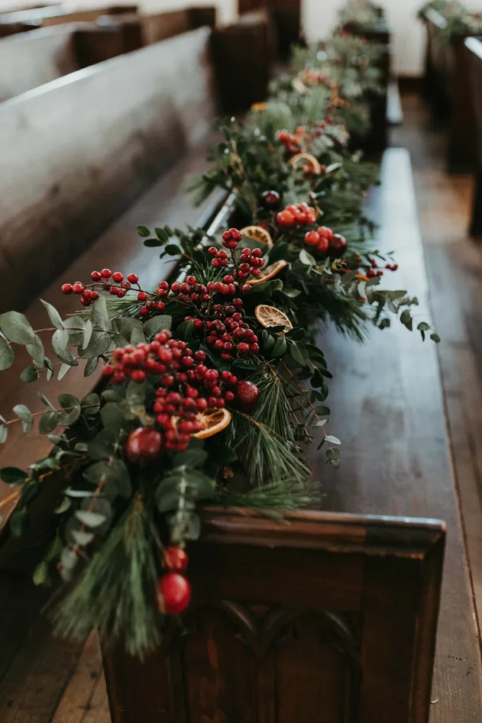 Garland of dark greenery and burgundy red berries draped along a wooden wedding pew