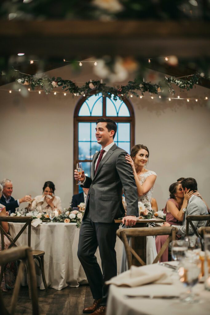  A man in a suit confidently holding a glass and speaking at a wedding reception, guests smiling in the background.