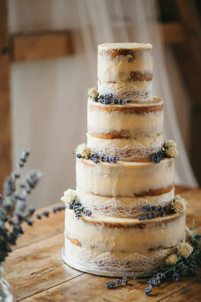 A three-tier cake with honey-toned buttercream (or honey cake showing through semi-naked frosting), delicate white lace trim or lace appliqué, fresh lavender sprigs tucked around, purple-grey and cream tones, romantic farmhouse.