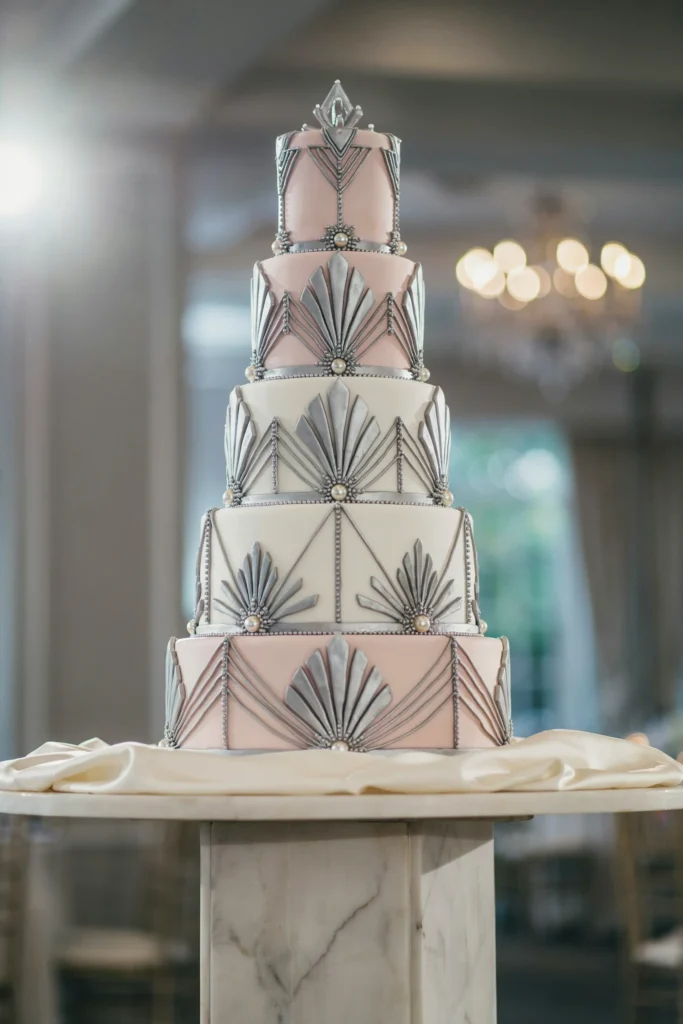 A four-tier cake, alternating blush and white tiers, platinum (silver-grey) geometric piping and accents, delicate linear patterns, stepped base like an Art Deco skyscraper, pearl or rhinestone accents at tier junctions.