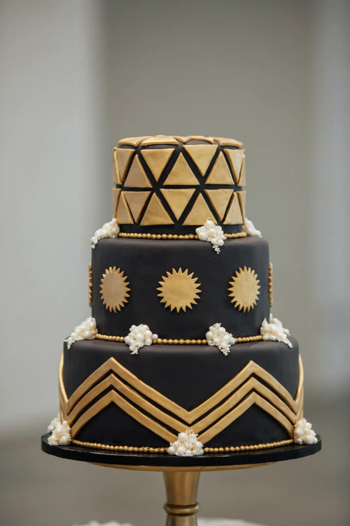 A three-tier square cake, black fondant base, bold gold geometric patterns (chevrons, sunbursts, triangles) piped on each tier, gold beaded borders, white sugar pearls clustered at corners, sleek and architectural.