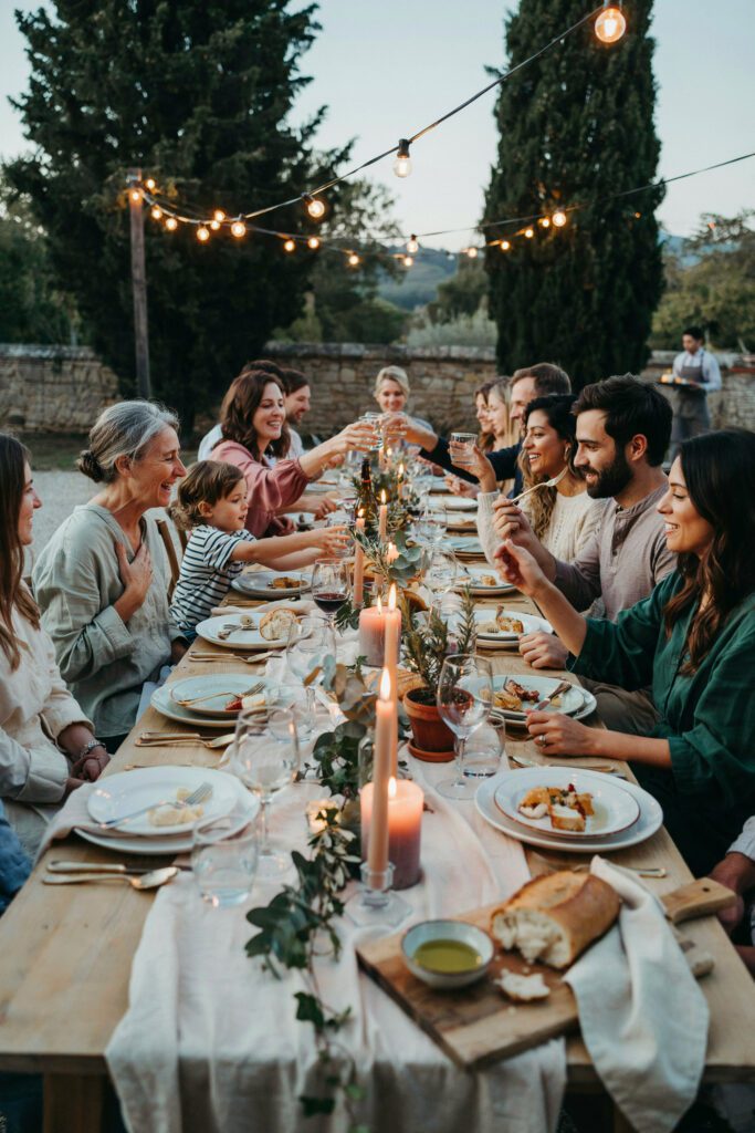 A long family-style dinner table at an intimate outdoor reception, decorated with candles, greenery, and simple place settings — guests laughing and eating together