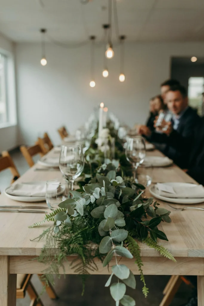 A low, wide bed of mixed greenery — eucalyptus, ferns, ruscus — running the length of a table with no tall elements blocking sightlines