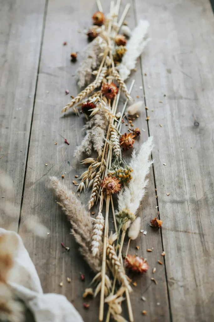 A loose, textural runner of dried wheat, pampas grass, and rust-toned dried flowers laid directly along a wooden table