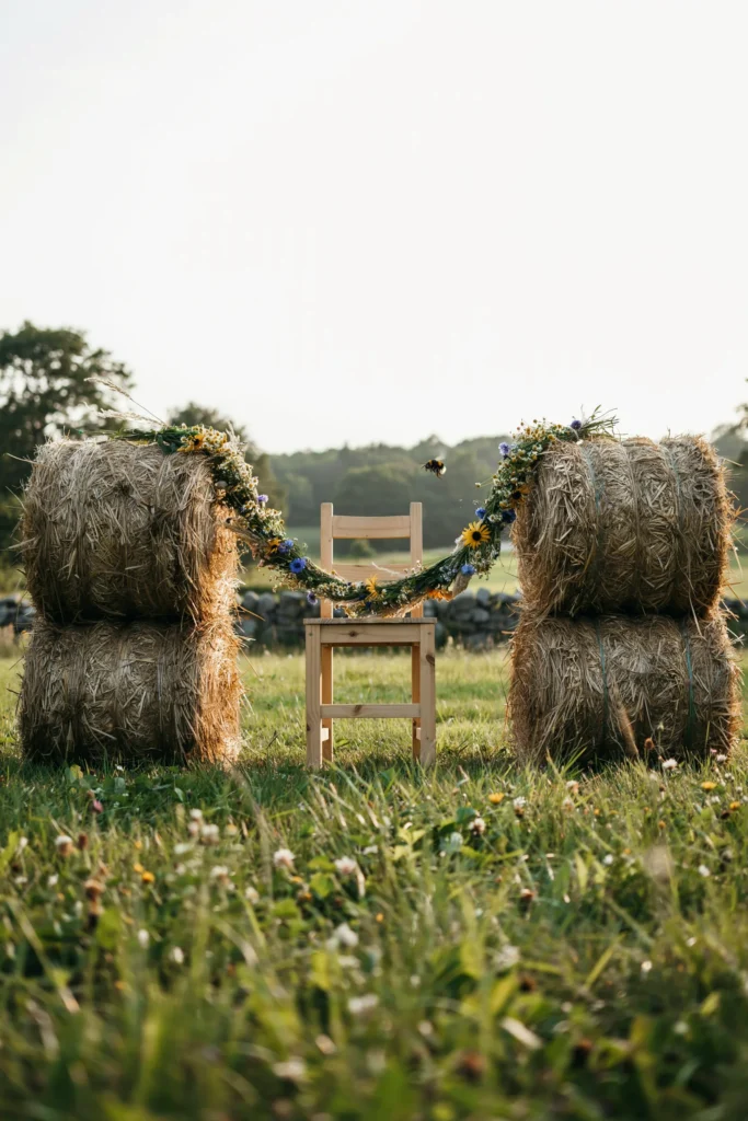 Two stacked hay bales flanking a simple wooden cross-back chair, wrapped in loose wildflower garland in warm yellows and creams