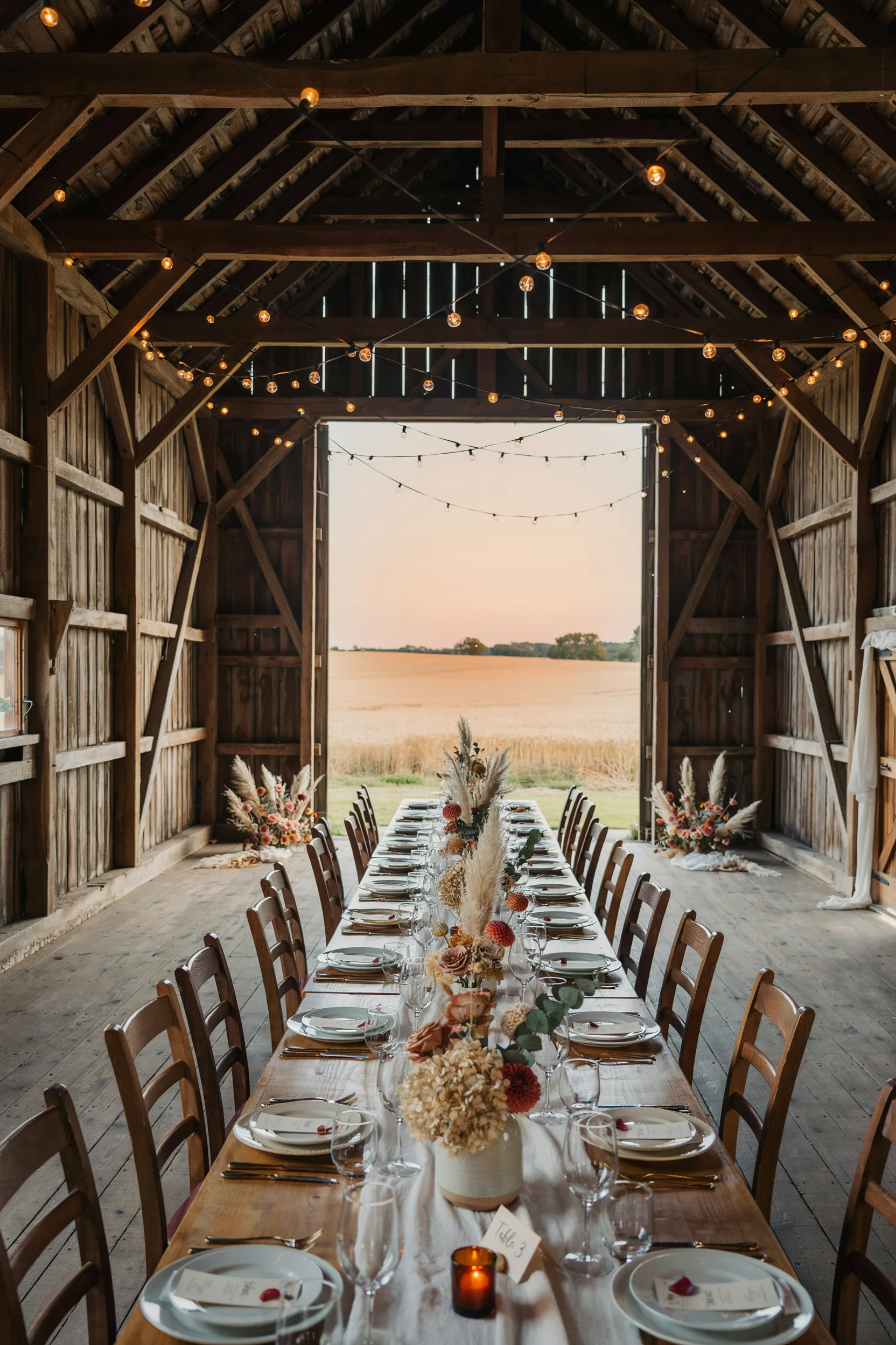 A softly lit barn reception at golden hour — string lights overhead, long wooden farm tables set with white linens and dried florals, doors open to a sunset field beyond.