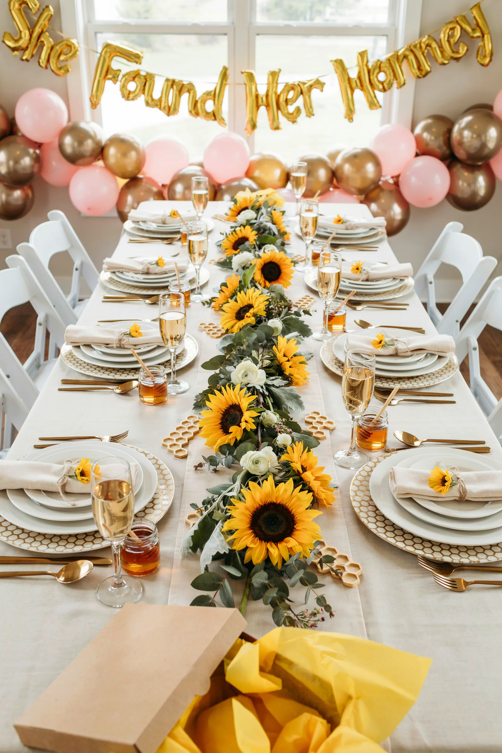 An overhead shot of a sun-drenched bridal shower table styled with sunflowers, honeycomb details, and gold flatware, with a "She Found Her Honey" balloon backdrop glowing in the background.