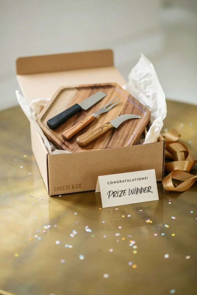 A small hexagon-shaped cheese board boxed gift set with mini cheese knives, styled as a game prize on a gold gift table.