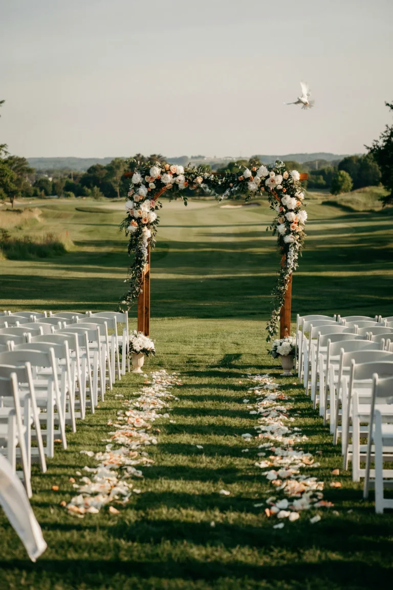 An elegant outdoor wedding ceremony set up on a manicured golf course fairway — white wooden chairs flanking a petal-strewn aisle, an arch of lush white and green florals at the far end, rolling greens in the background under golden afternoon light.