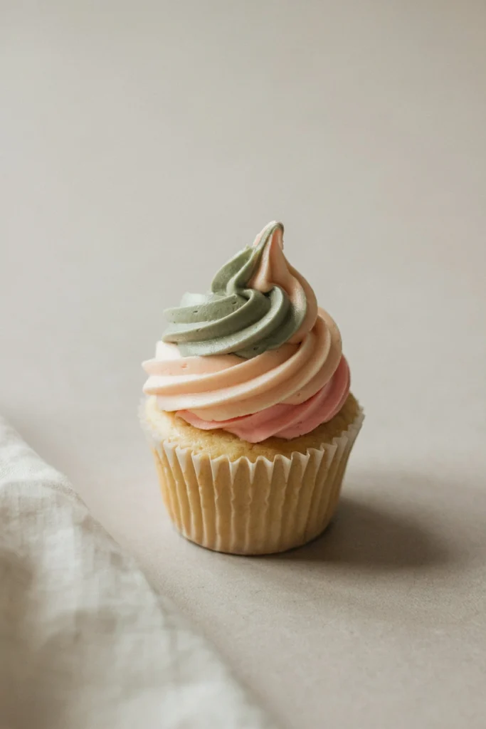 A cupcake with a tall swirl of hand-painted watercolor buttercream blending from dusty sage at the base to soft blush pink at the tip, with no additional decoration, the gradient doing all the work