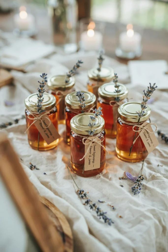 Small glass honey jars with gold lids, each tied with a sprig of dried lavender and a card printed with "Sweet beginnings — [Couple's names]." The jars are clustered on a wooden cutting board surrounded by scattered dried rose petals.