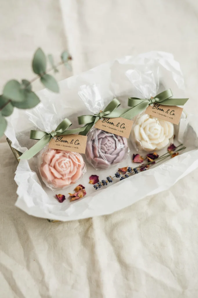 Small bars of flower-shaped soap in blush, lavender, and white, wrapped in clear cellophane tied with a sage ribbon and a small card. Each soap is pressed in the shape of a rose or peony bloom. They're displayed in a shallow tray lined with white tissue paper.
