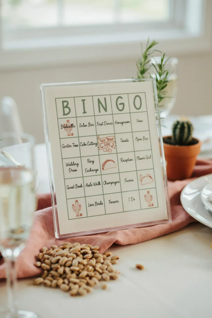 A custom bingo card for a bridal shower printed on cream cardstock with small cactus and taco illustrations in each corner — the center free square reads "Taco 'Bout Love" — displayed in a small acrylic frame at each place setting alongside a small pile of dried pinto beans as markers.