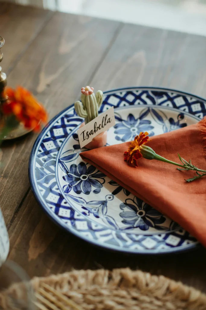  A close-up of a single place setting featuring a cobalt blue and white Talavera-style patterned plate, a terracotta linen napkin folded into a triangle, a single stem orange marigold laid across the napkin, and a small hand-lettered place card standing in a tiny cactus-shaped card holder.