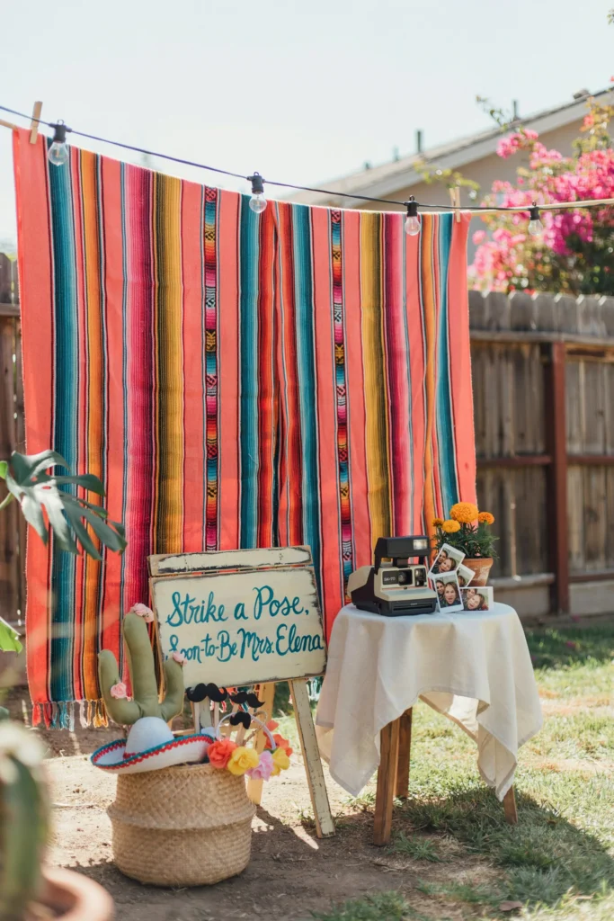 A DIY photo booth corner with a bright serape fabric backdrop, a basket of handmade props (felt cactus on sticks, mini sombrero headbands, mustache on a stick), a polaroid camera on a small table, and a sign reading "Strike a Pose, Soon-to-Be Mrs. [Name]" in chalk lettering.
