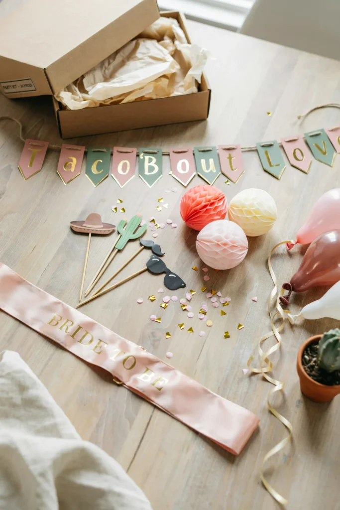 An unboxed bridal shower party kit displayed across a table — banner letters spelling "Taco 'Bout Love," honeycomb tissue balls, a bride-to-be sash, photo booth props including a sombrero cutout and cactus frame, and a set of coordinating confetti balloons.