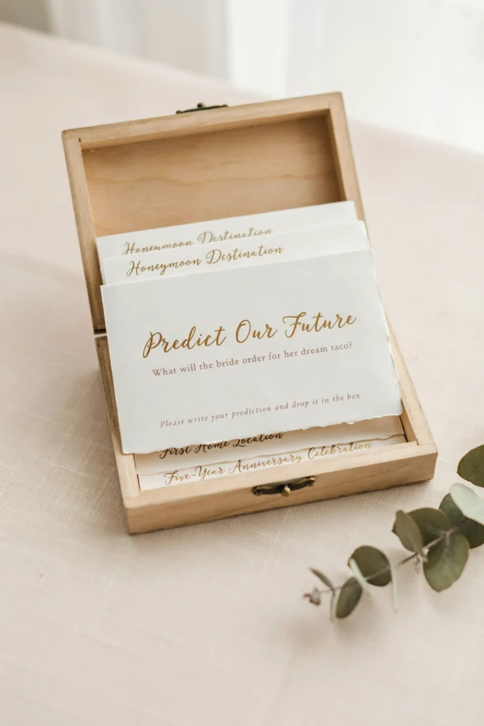 Custom printed prediction cards asking guests to predict the bride's dream taco order, honeymoon destination, first home location, and five-year anniversary celebration — styled in dusty rose and gold script, displayed in a small wooden card box.]