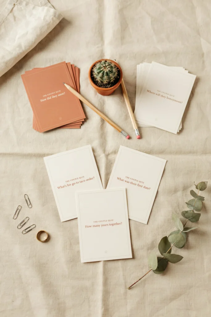 A styled game card flat lay — printed trivia cards in terracotta and cream asking questions like "How did they meet?", "What's his go-to taco order?", and "Where will they honeymoon?", laid out with mini pencils and a small cactus centerpiece.