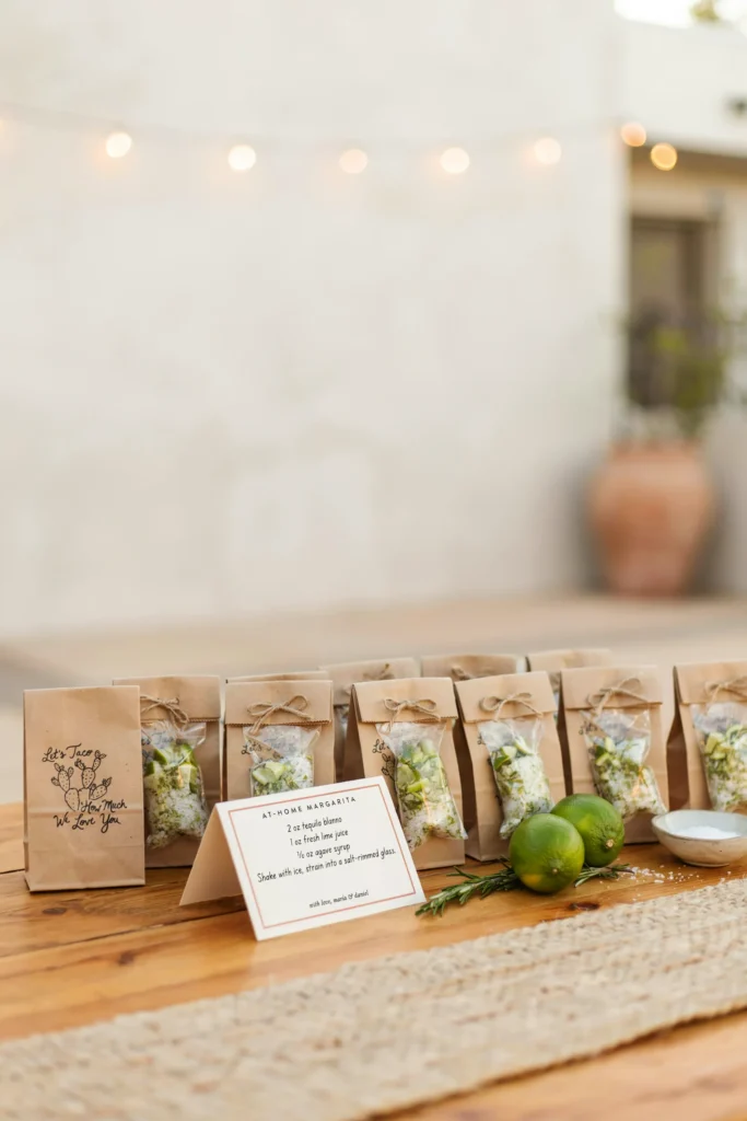 A styled favor table with small kraft bags filled with individual margarita mix packets, each bag stamped with a lime and cactus illustration and the text "Let's Taco 'Bout How Much We Love You," displayed alongside a small card with a simple at-home margarita recipe.