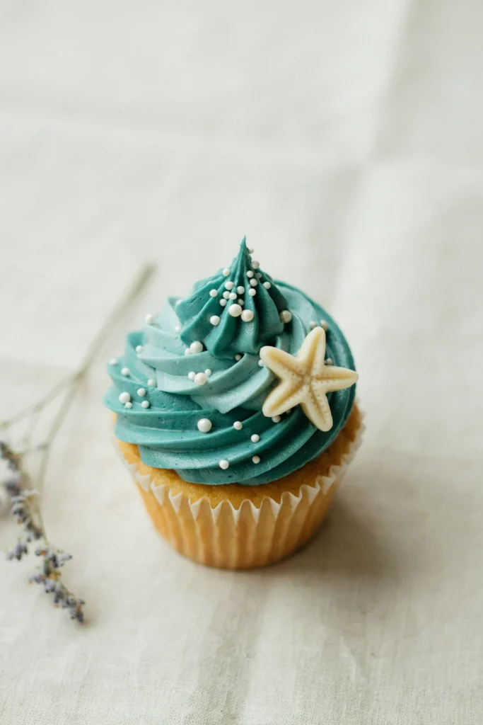 A cupcake with swirled sea glass blue-green buttercream, the surface scattered with irregular white pearl sugar beads at various sizes, a single tiny fondant starfish pressed into one side of the swirl