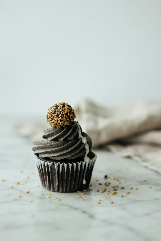 A cupcake with a swirl of deep charcoal-grey black sesame buttercream, a thin white ganache drip running down one side, and a single sesame-coated chocolate truffle balanced on top