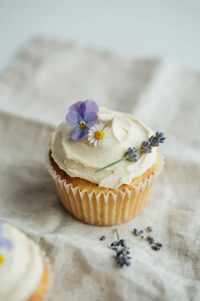 A cupcake with soft cream cheese frosting smoothed loosely to the edges, topped with three tiny fresh flowers — one violet pansy, one micro daisy, and one sprig of lavender — arranged asymmetrically