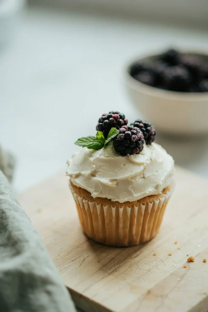 A cupcake with rough, palette-knife-textured frosting in ivory — the surface deliberately uneven, showing ridges and peaks — topped with a small cluster of fresh blackberries and two green mint leaves