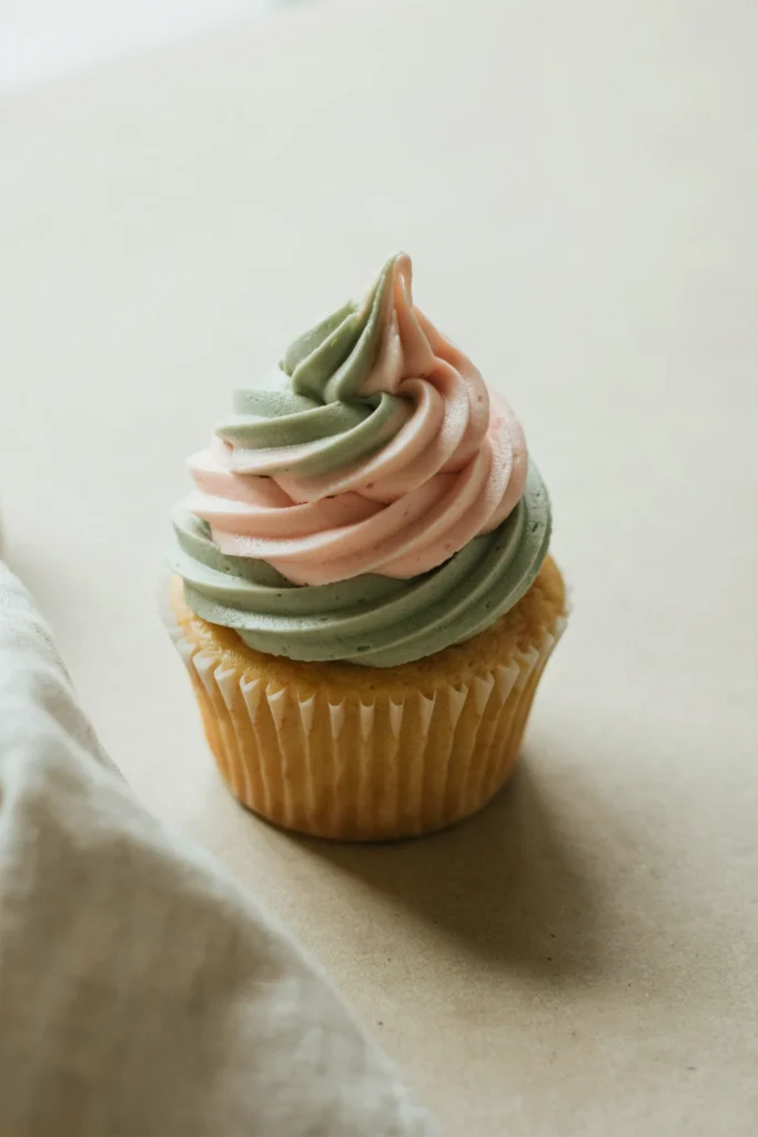 A cupcake with a smooth amber-toned honey cream cheese frosting, topped with a single pressed dried wildflower — deep burgundy and cream petals fanned flat — with a very thin glaze that gives the whole top a translucent sheen