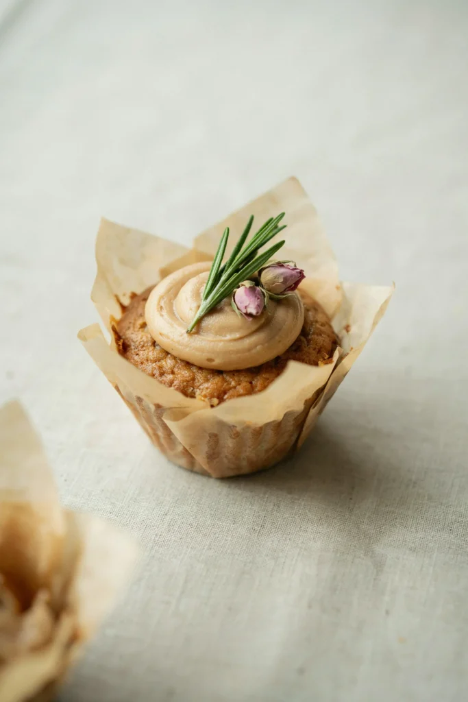 A cupcake on a natural kraft paper liner with barely-there buttercream leaving the edges of the cake visible, topped with a single fresh rosemary sprig and two tiny dried rosebuds