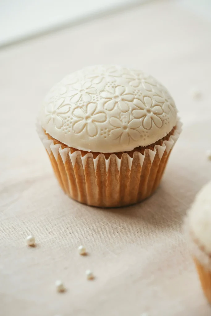 A flat-topped cupcake covered in smooth white fondant with an intricate lace impression pressed into the surface, sitting on a champagne-colored scalloped liner