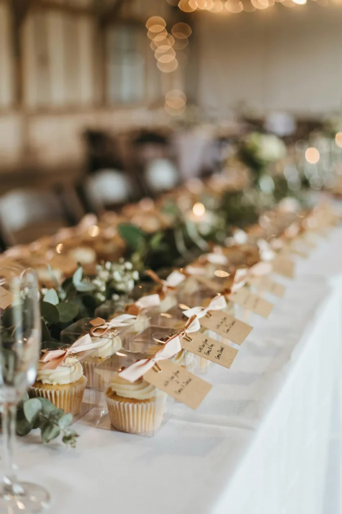 A display of plain frosted cupcakes sitting inside individual clear acetate favor boxes tied with satin ribbon, displayed in rows on a table with a small handwritten tag on each reading "With Love from [Couple's Name