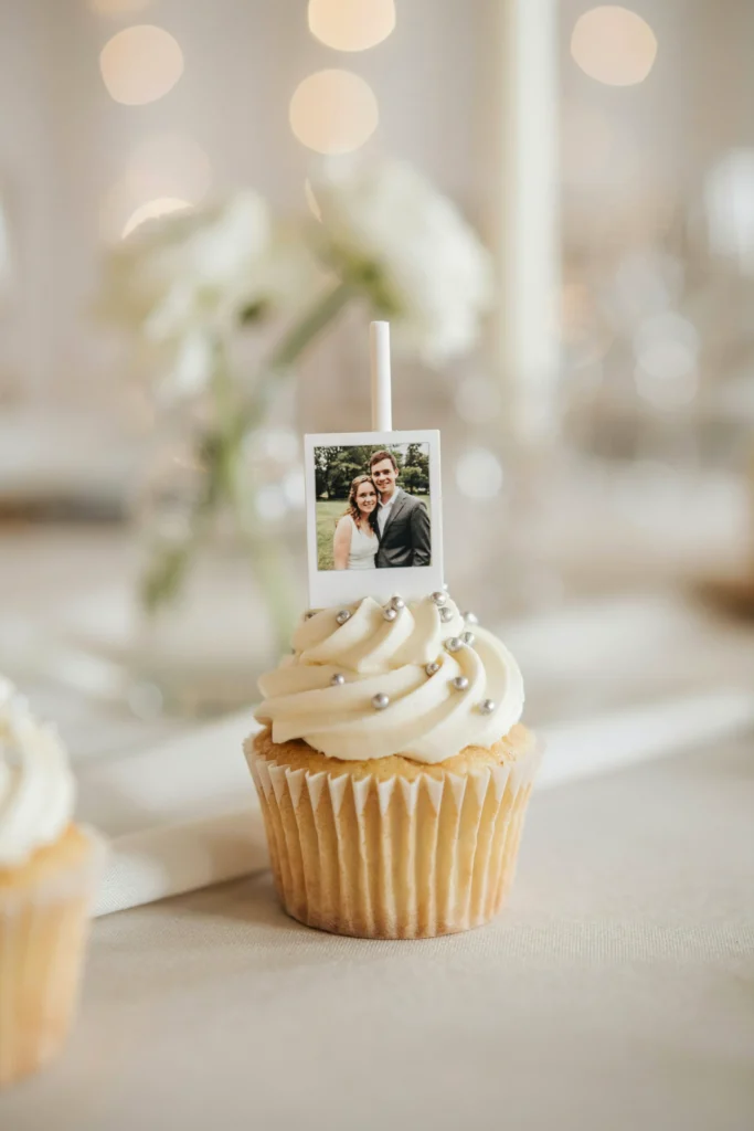 A cupcake with white buttercream and a tiny edible polaroid-style photo printed on wafer paper — a miniature image of the couple — mounted on a thin white pick and placed upright in the frosting