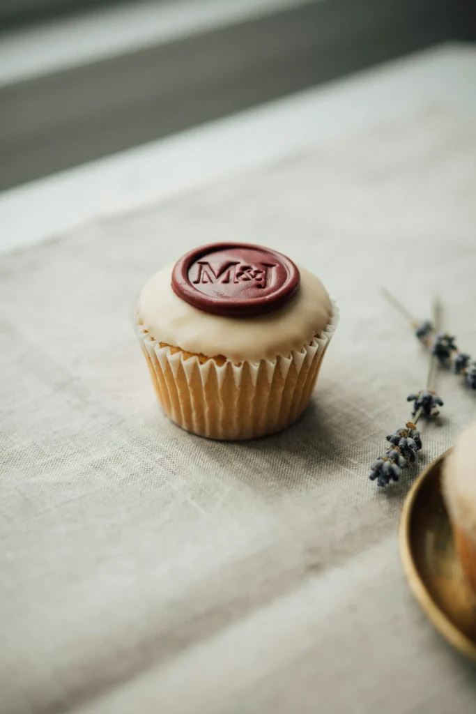 A cupcake with a smooth cream fondant top bearing a circular fondant disc in deep burgundy that mimics a wax seal — with the couple's initials pressed into it — sitting slightly off-center on the frosting